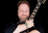 Mitch Benn holding a guitar and smiling against a black background Mitch Benn holding a guitar and smiling against a black background