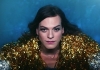 A woman in a gold and silver sparkly top, wearing false eyelashes on a blue background looks into the camera A woman in a gold and silver sparkly top, wearing false eyelashes on a blue background looks into the camera