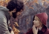 A woman with short blonde hair, wearing a red coat, talks to a man with curly, dark hair and glasses outside on a Autumn day. A woman with short blonde hair, wearing a red coat, talks to a man with curly, dark hair and glasses outside on a Autumn day.
