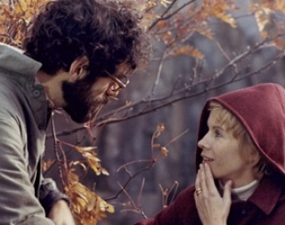A woman with short blonde hair, wearing a red coat, talks to a man with curly, dark hair and glasses outside on a Autumn day. A woman with short blonde hair, wearing a red coat, talks to a man with curly, dark hair and glasses outside on a Autumn day.