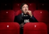 Comedian Andrew Maxwell dressed in black sitting on red cinema seats eating popcorn Comedian Andrew Maxwell dressed in black sitting on red cinema seats eating popcorn