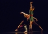 Three dancers perform on a black background and floor, dressed in bright colours Three dancers perform on a black background and floor, dressed in bright colours