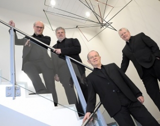 Four musicians from the Coull Quartet stand on a glass and chrome staircase, wearing black suits Four musicians from the Coull Quartet stand on a glass and chrome staircase, wearing black suits