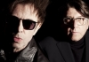 Two men in black clothes, wearing glasses, pose against a black background Two men in black clothes, wearing glasses, pose against a black background