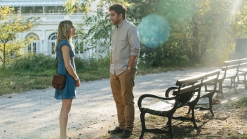 A woman in a blue dress talks to a man in a grey shirt, standing in front of a park bench in the sun. A woman in a blue dress talks to a man in a grey shirt, standing in front of a park bench in the sun.