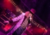 A man with a large beard and a top hat speaks into a mic, lit by purple lighting A man with a large beard and a top hat speaks into a mic, lit by purple lighting