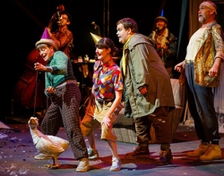 Four performers, two female and two male, in colourful clothes, stand in a line behind a puppet of a white duck Four performers, two female and two male, in colourful clothes, stand in a line behind a puppet of a white duck