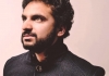 Nish Kumar wearing a black jacket standing against a pale pink background. Nish Kumar wearing a black jacket standing against a pale pink background.