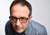 A man wearing a blue, striped shirt and black rimmed glasses leans towards the camera A man wearing a blue, striped shirt and black rimmed glasses leans towards the camera